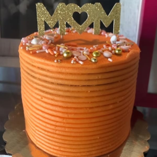 Orange frosted cake with sprinkles and a gold glitter "MOM" topper.