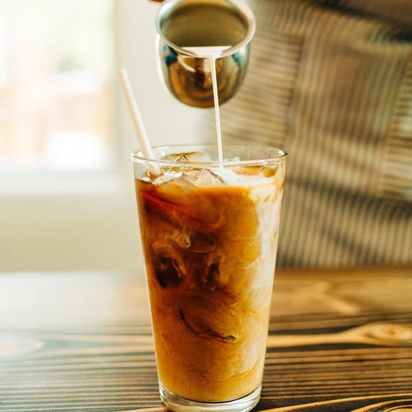 Cream being poured into an iced coffee with a straw on a wooden table.