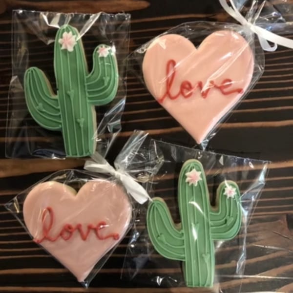 Four individually wrapped cookies, two green cacti with pink flowers, and two pink hearts with "love" written in red.