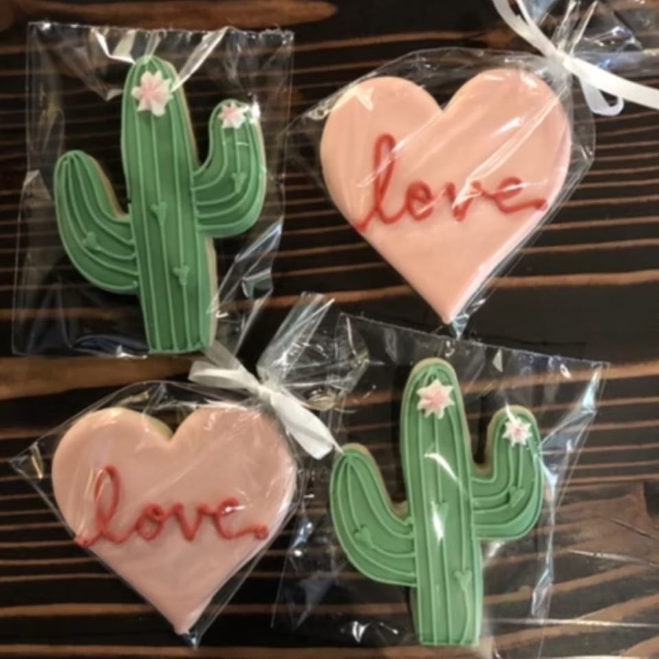 Four individually wrapped cookies, two green cacti with pink flowers, and two pink hearts with "love" written in red.