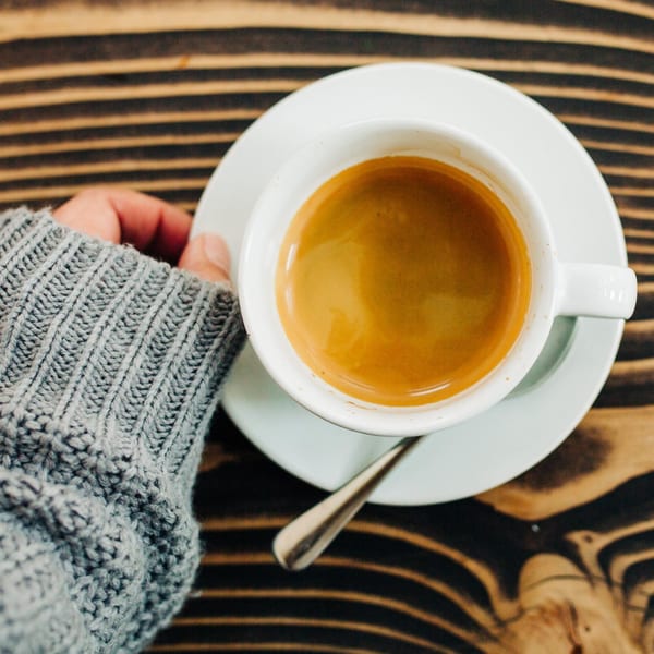 Overhead view of a hand in a gray sweater holding a white cup of espresso on a wooden table.