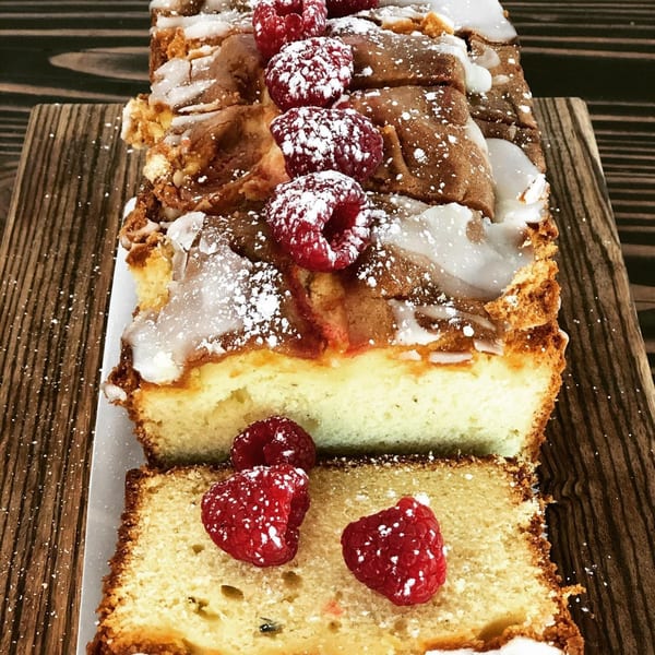 Raspberry loaf cake with white icing and powdered sugar, with a slice cut, on a wooden board.