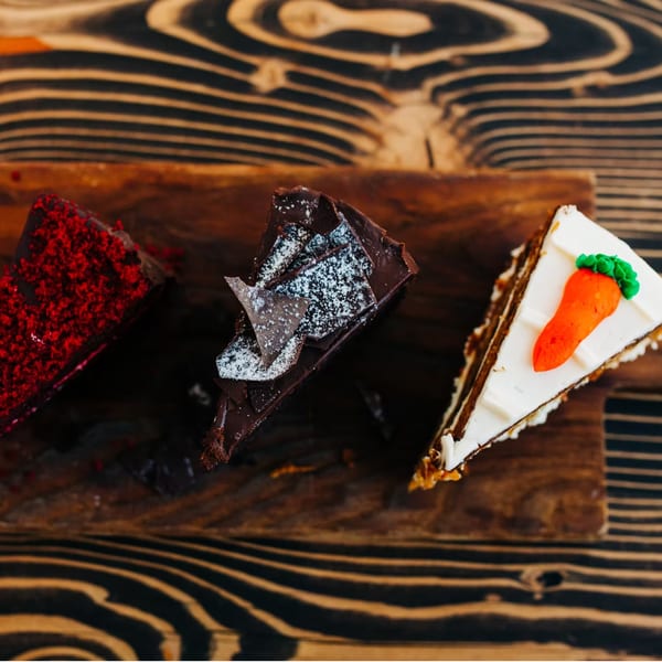 Three slices of cake: red velvet, chocolate, and carrot cake, on a wooden board.
