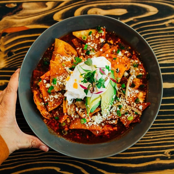 A hand holds a bowl of Chula Artisan Eatery's chilaquiles with avocado, poached egg, and cheese.