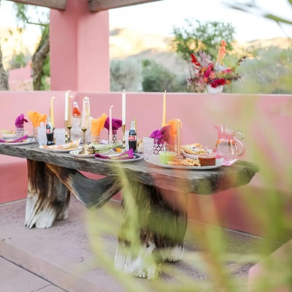 Outdoor dining table set with Mexican food, drinks, candles, and colorful napkins against a pink wall.