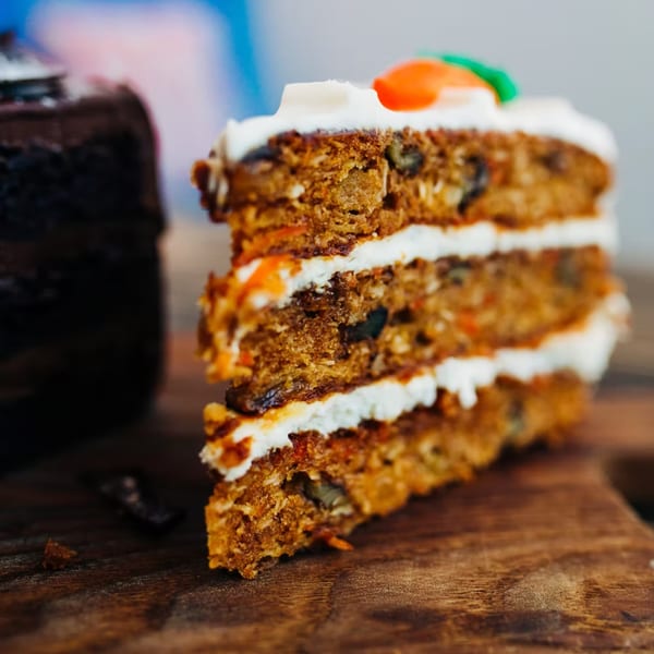 A slice of carrot cake with white frosting and a chocolate cake in the background.