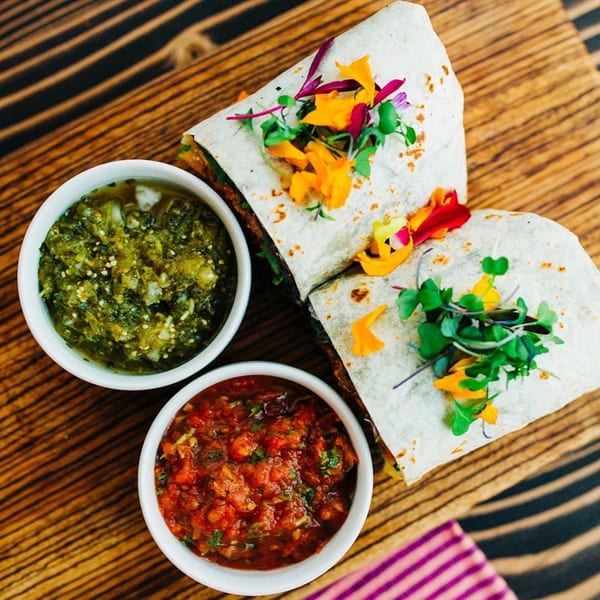 Two Chula Artisan Eatery burritos with edible flowers and microgreens, served with green and red salsa.