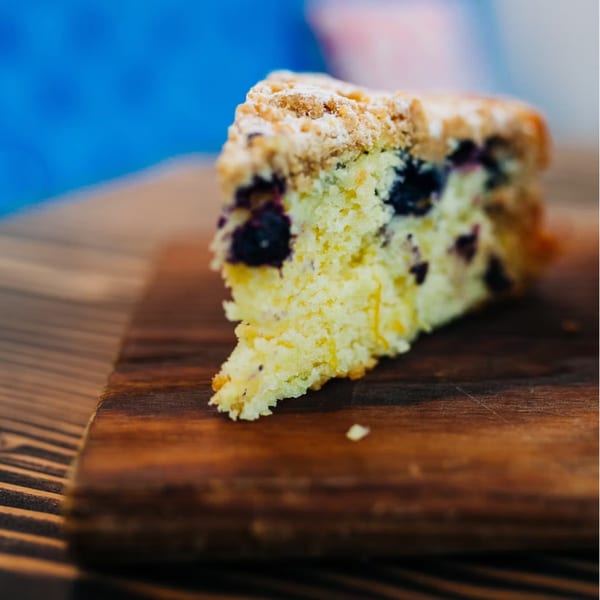 A slice of blueberry crumble cake with powdered sugar on a wooden board.