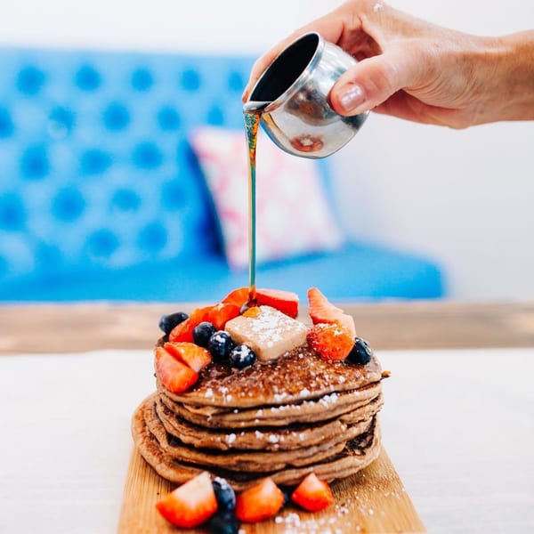 Syrup being poured onto a stack of pancakes with strawberries, blueberries, and butter.