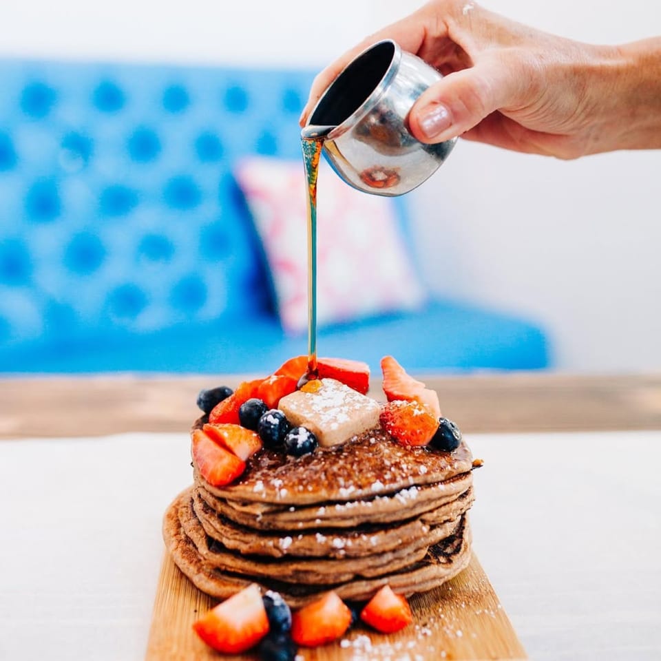 Syrup being poured onto a stack of pancakes with strawberries, blueberries, and butter.