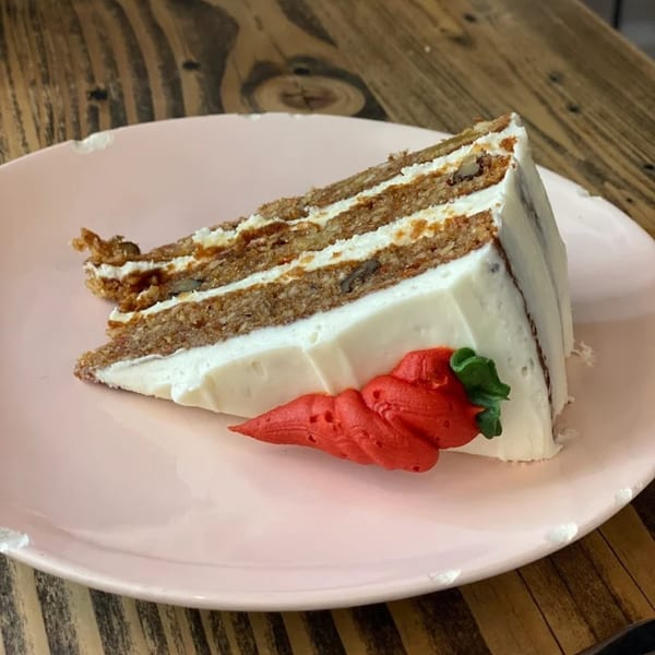 A slice of carrot cake with white frosting and a red carrot decoration on a pink plate.