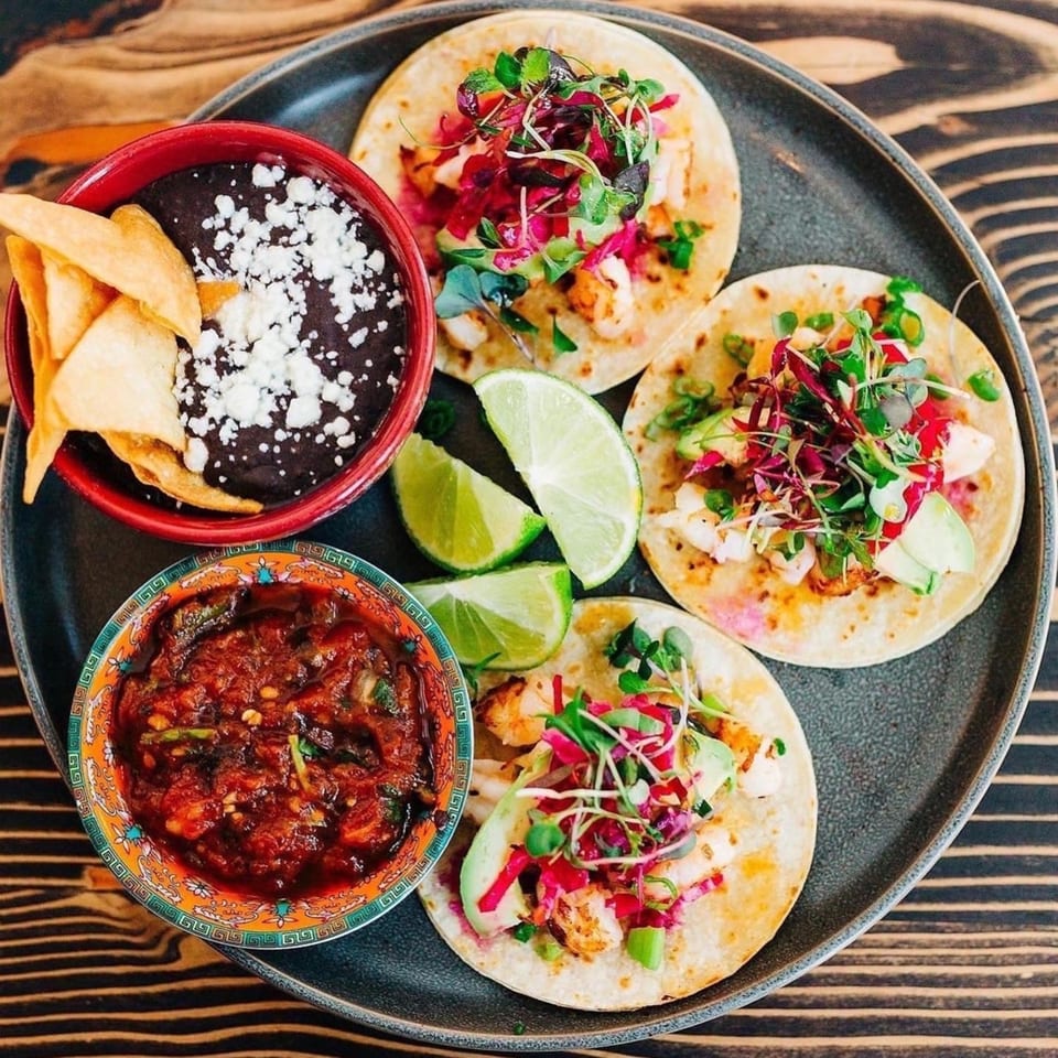 Three Chula Artisan Eatery tacos with sides of black beans, salsa, and lime wedges.