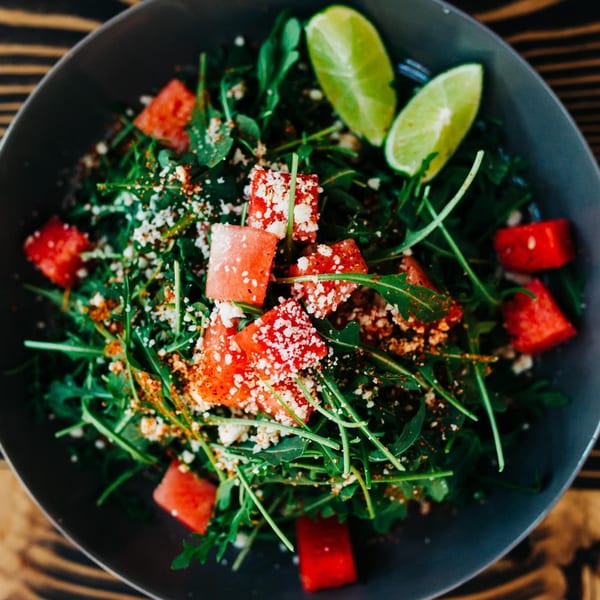 A vibrant Chula Artisan Eatery salad with arugula, watermelon, cotija cheese, and lime wedges.