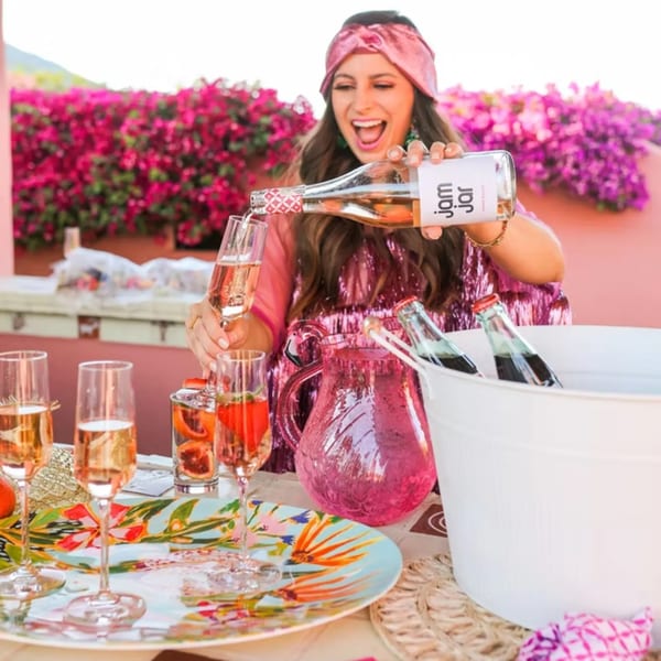 A woman in a pink sequin dress pours rosé wine into a glass on a vibrant patio.